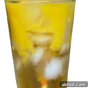 A tall glass filled with iced Mango Greentini, featuring a yellow layer on top and a pink-striped straw. The text Mango Greentini and a small mango illustration appear above the drink.
