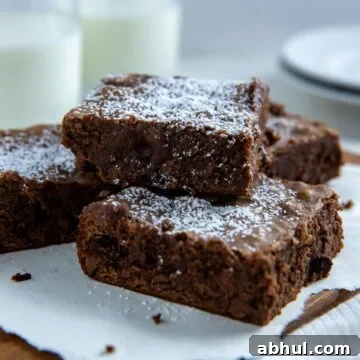 Four baked brownies sprinkled with powdered sugar on a wooden platter with two glasses of milk in the background.