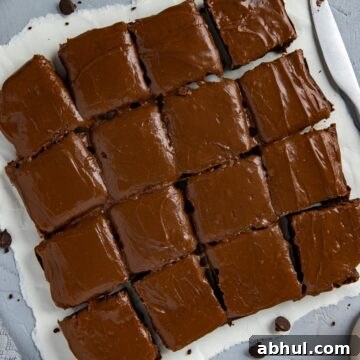 Baked and frosted brownies cut into 16 squares on white parchment paper with brownie crumbs scattered around.