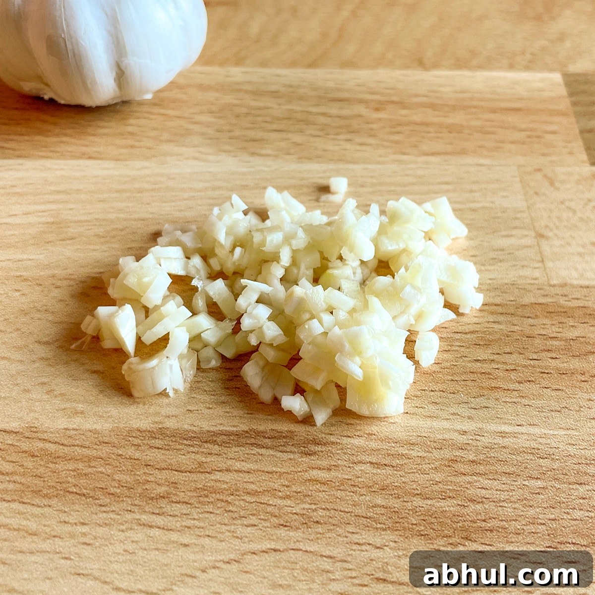 Minced garlic with a head of garlic in the background on a butcher block cutting board