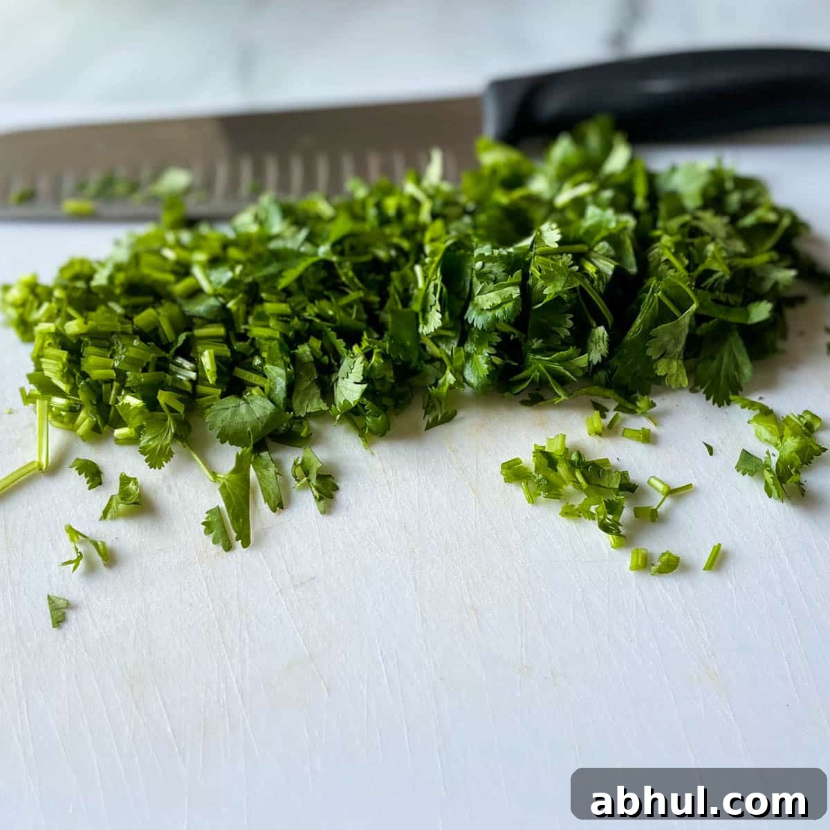 Chopped fresh cilantro is shown on a white cutting board with a kitchen knife in the background.