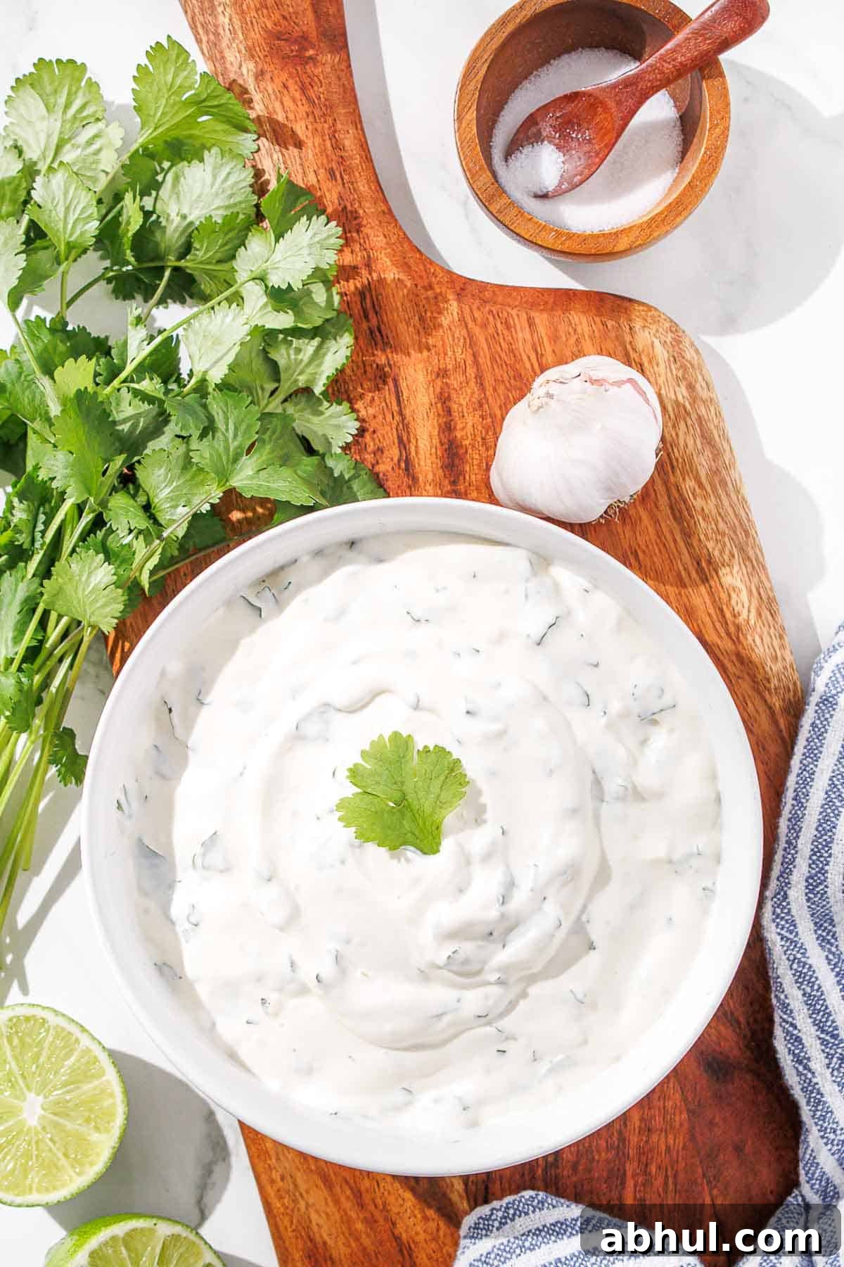 A bowl of cilantro lime crema on a wooden board, flanked by fresh cilantro, lime, garlic, and a pinch bowl of salt.