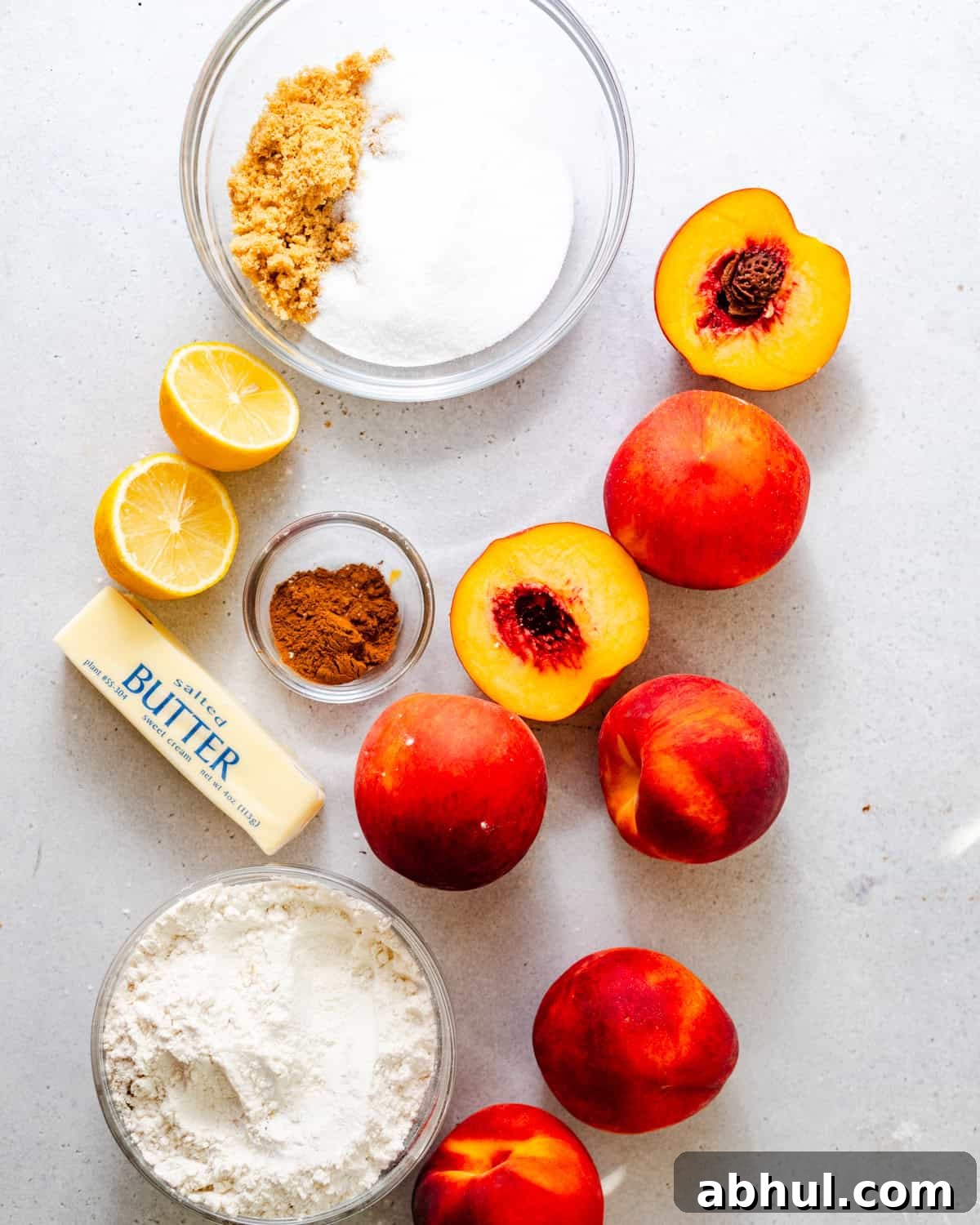 ingredients to make peach crumble on a white surface - fresh peaches, spices, brown sugar, flour, cornstarch, butter, and lemon juice.