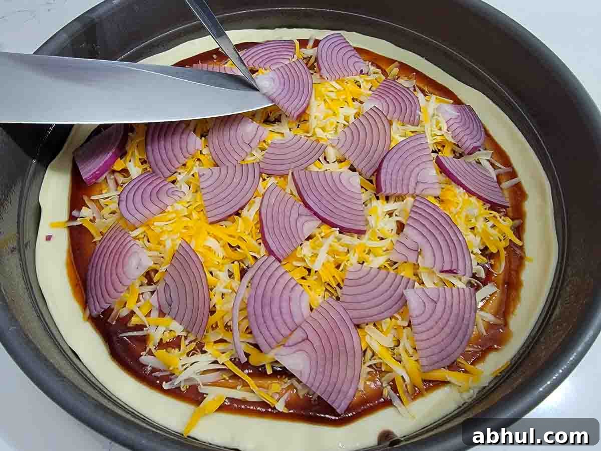 red onions slices and shredded cheese added to pizza dough and sauce in a pan.