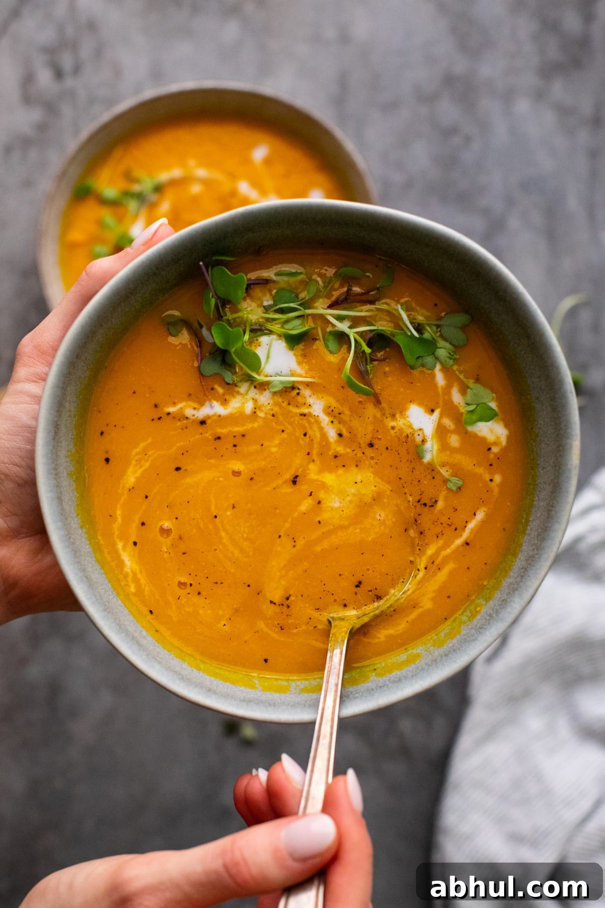 hands holding a gray bowl filled with orange carrot ginger soup with a hand holding a soup in it.