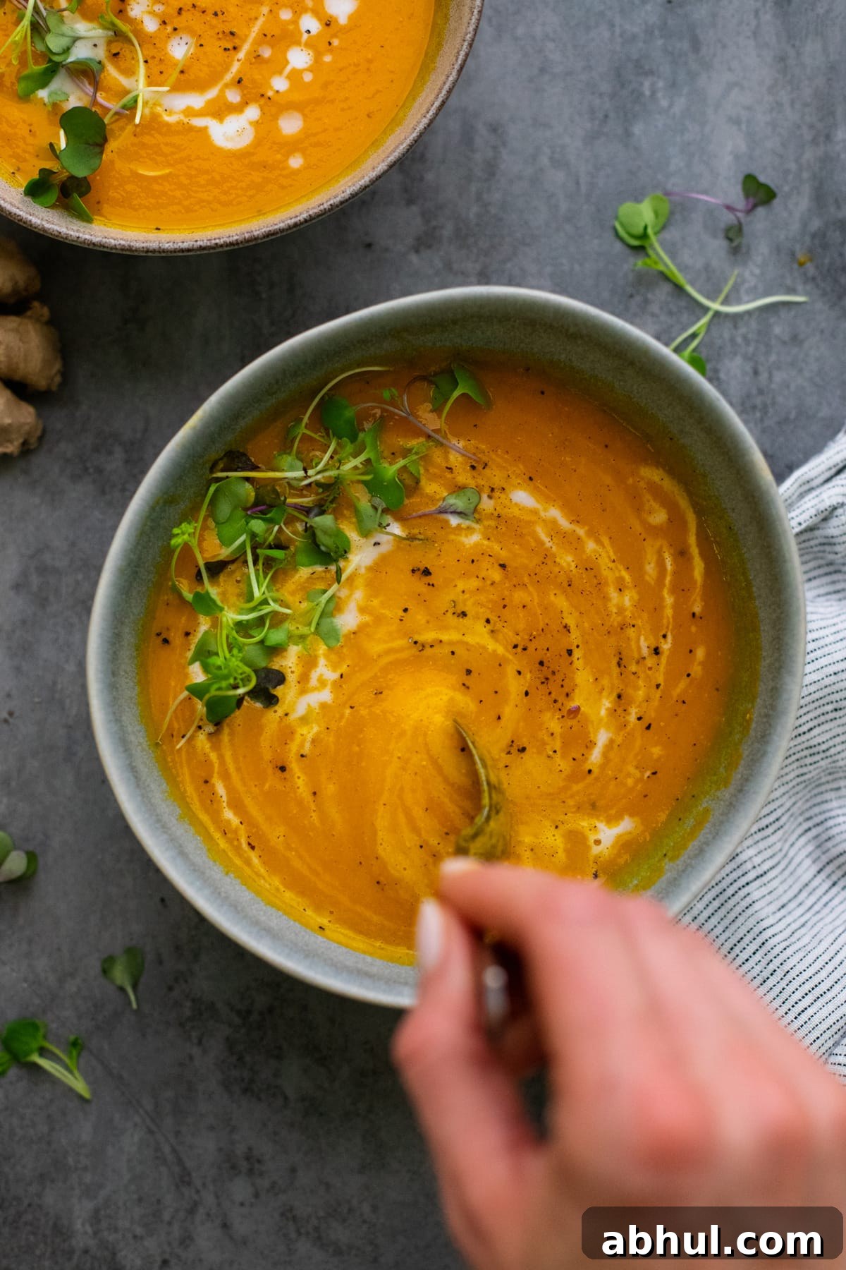 hand using spoon to stir orange carrot ginger soup in ceramic bowl on gray background.