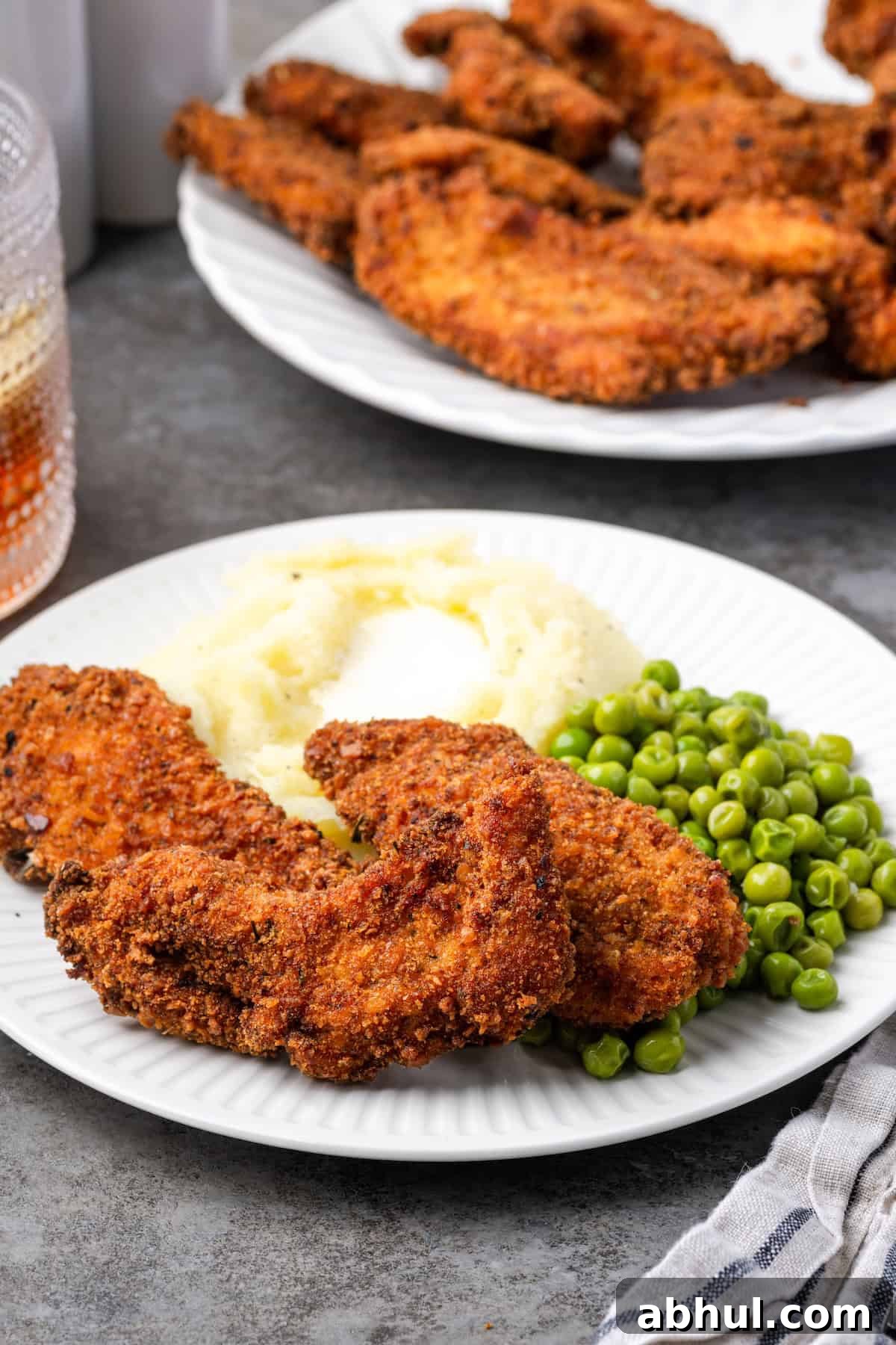 Crispy chicken tenders breaded with chicken stuffing next to mashed potatoes and peas on a plate, with a platter of chicken tenders in the background.