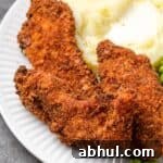 Close up of crispy chicken tenders breaded with chicken stuffing next to mashed potatoes and peas on a plate.