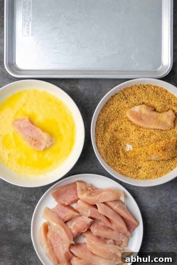 Overhead view of a chicken breading station with a bowl of chicken, stuffing crumbs, and an egg wash next to a baking pan.