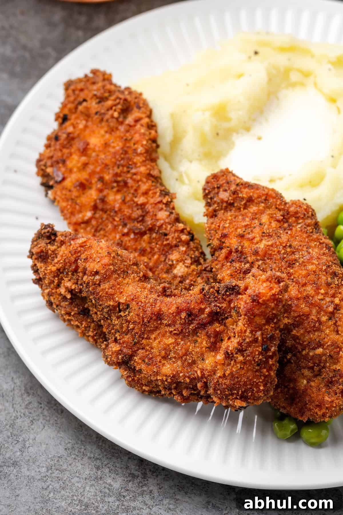 Close up of crispy chicken tenders breaded with chicken stuffing next to mashed potatoes and peas on a plate.