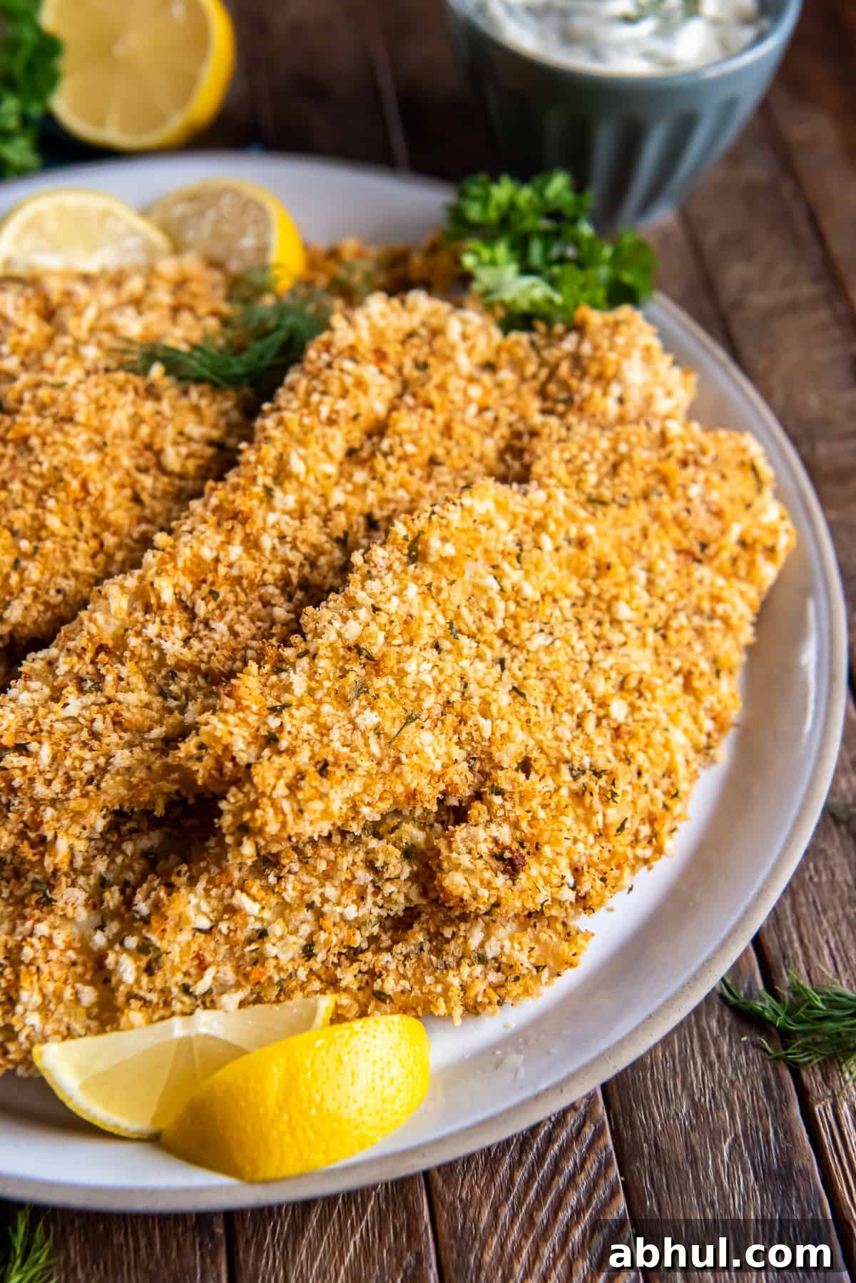 Crispy Oven-Baked Fish Fillets with Parmesan Crust 9 A close up of crispy oven baked fish on a white plate with lemon wedges and fresh dill.