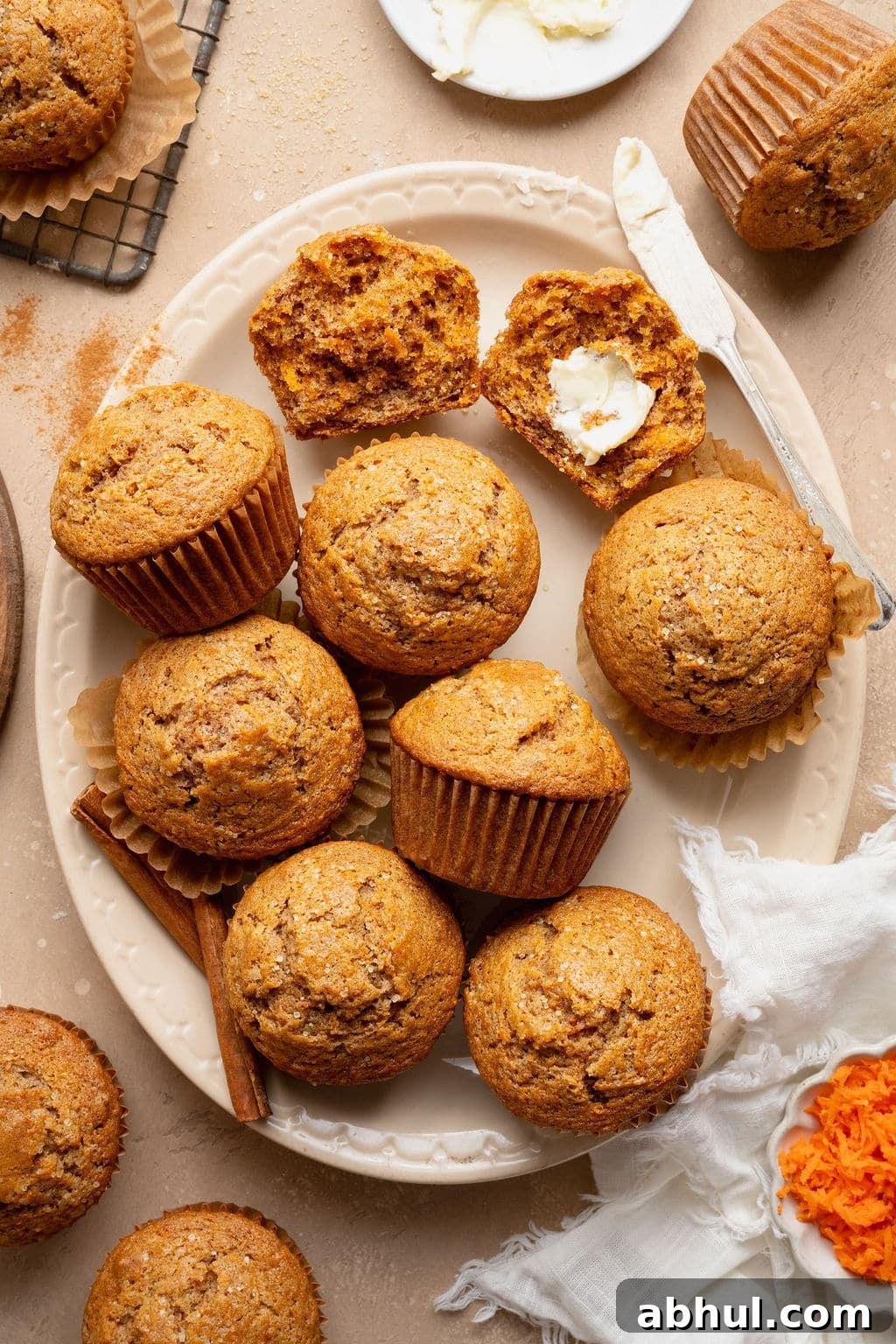Platter of carrot cake muffins with one split open and spread with butter, surrounded by cinnamon sticks, grated carrots, and a butter knife.