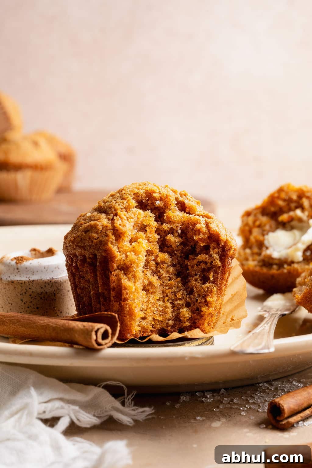 Single carrot cake muffin on a plate with cinnamon sticks and a small jar of cinnamon in the background, styled with soft neutral linens.