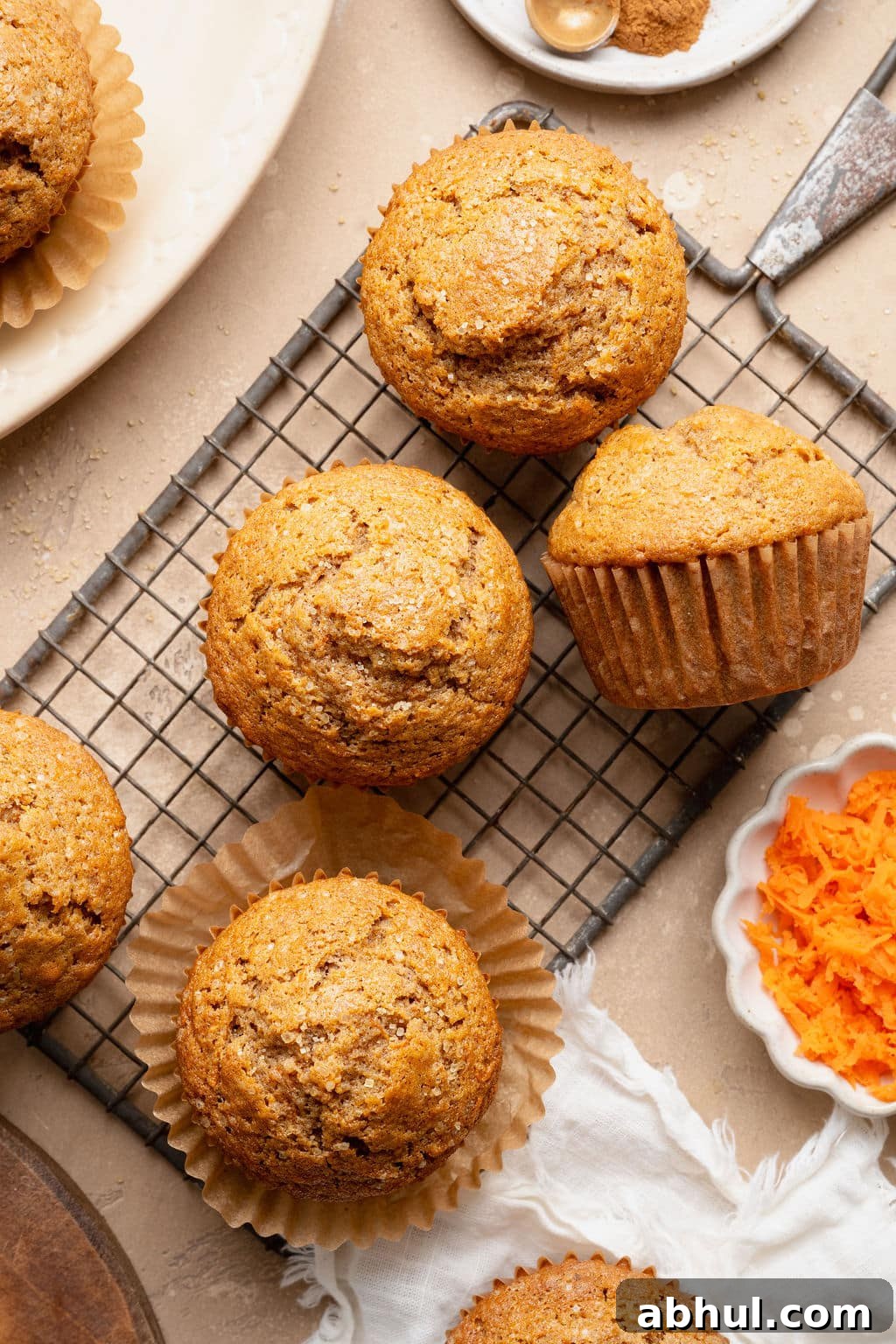 Overhead view of carrot cake muffins cooling on a wire rack, topped with coarse sugar, with grated carrots and cinnamon nearby on a neutral background.
