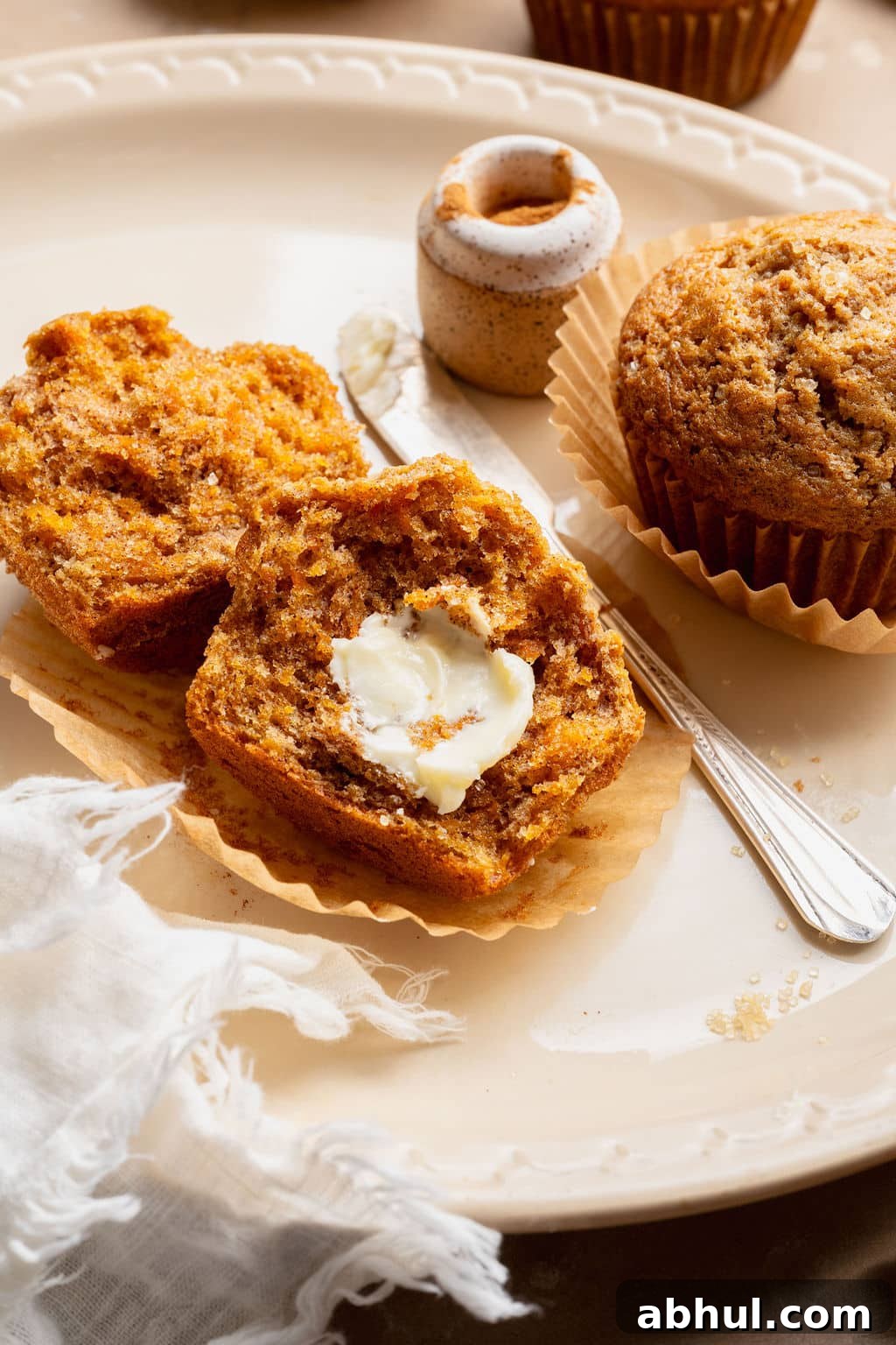 Carrot cake muffin broken in half and topped with softened butter on a parchment liner, with a butter knife and cinnamon jar nearby.
