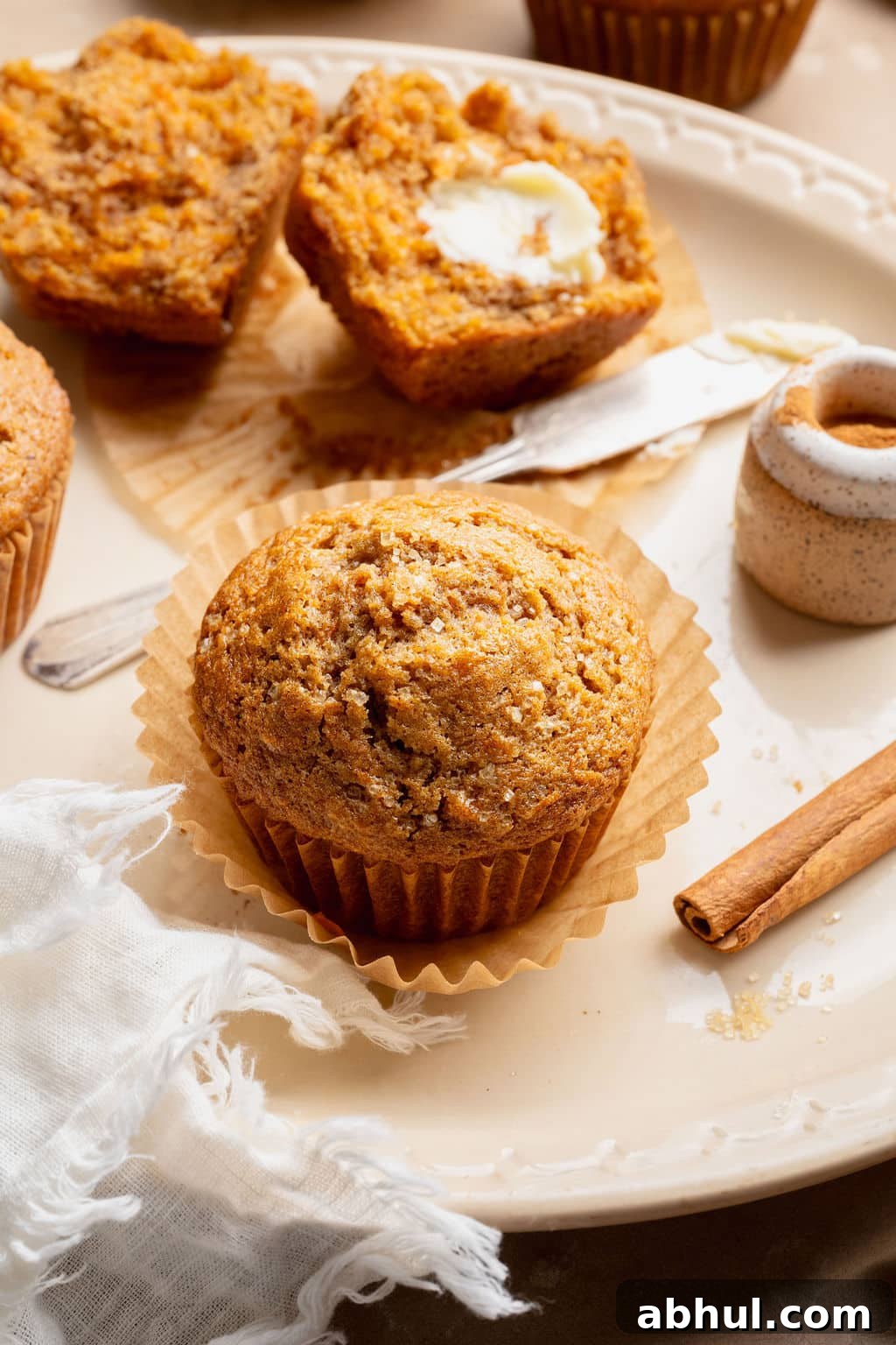 Single carrot cake muffin on a plate with cinnamon sticks and a split muffin in the background, highlighting the domed top and sugar crust.