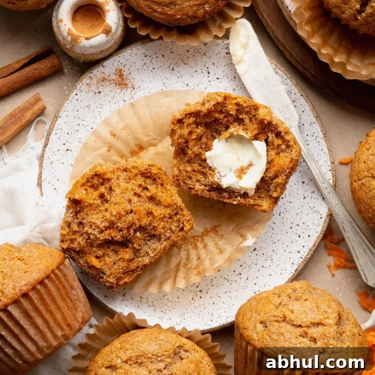 Single carrot cake muffin on a plate with cinnamon sticks and a split muffin in the background, highlighting the domed top and sugar crust.
