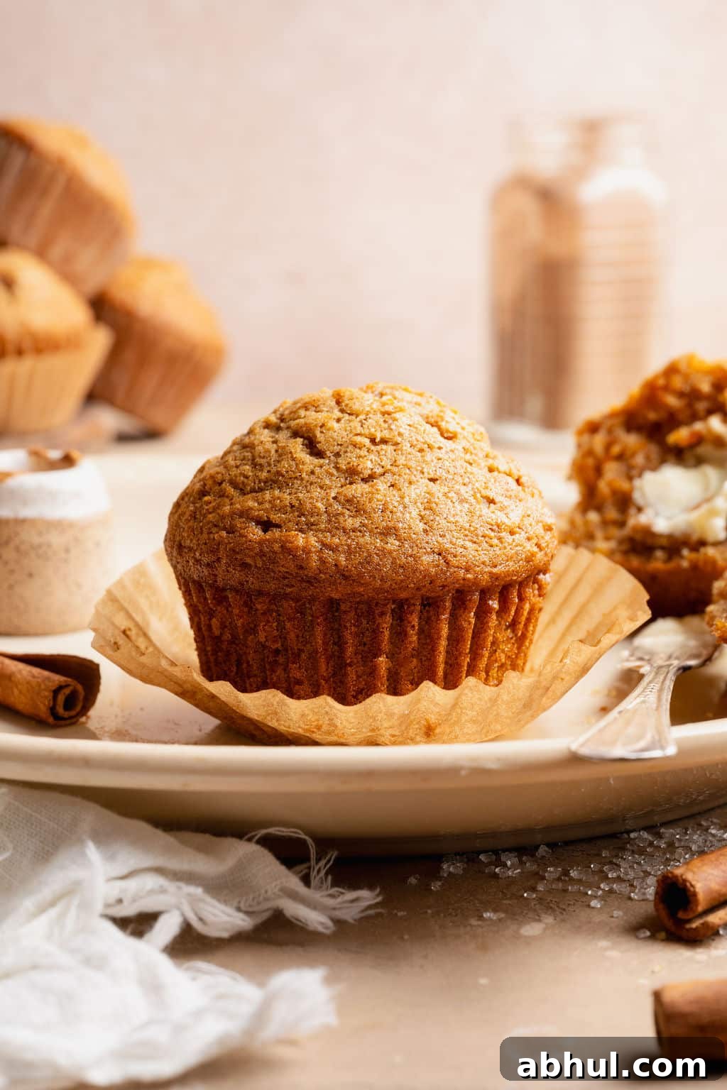 Side view of a carrot cake muffin in a paper liner on a plate, with another muffin split open and buttered in the background.