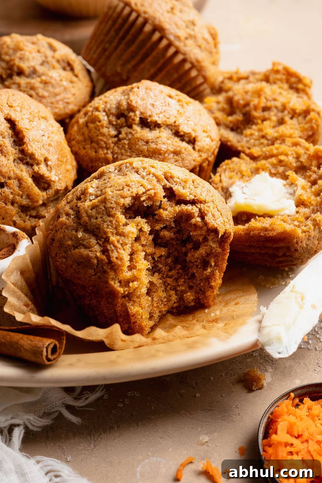 Close-up of a carrot cake muffin with a bite taken out, showing a moist, tender crumb, served on a plate with butter and cinnamon sticks.