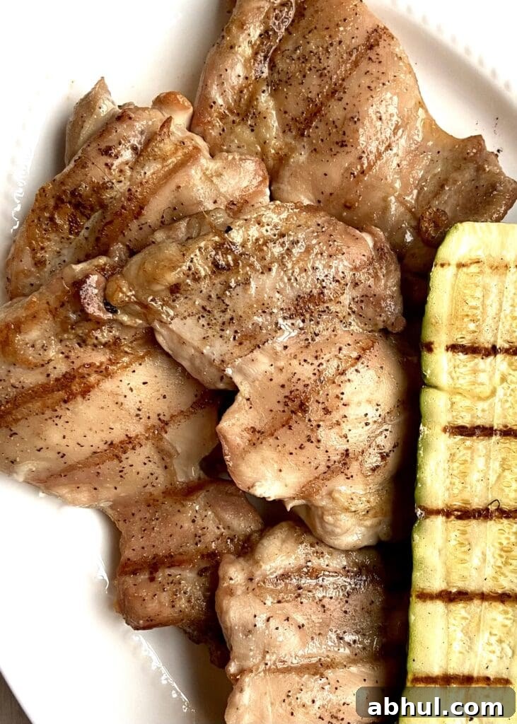 Close up of grilled boneless skinless chicken thighs on a white platter next to grilled zucchini.