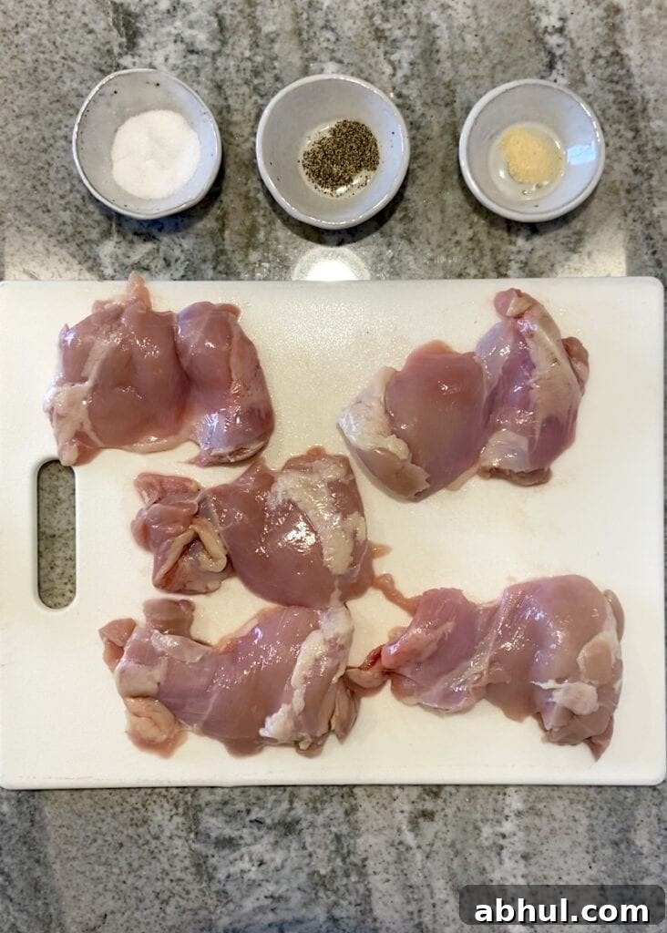 Top down shot of ingredients for grilled boneless skinless chicken thighs, including 5 chicken thighs on a white cutting board and small gray bowls with salt, pepper, and garlic powder in them.