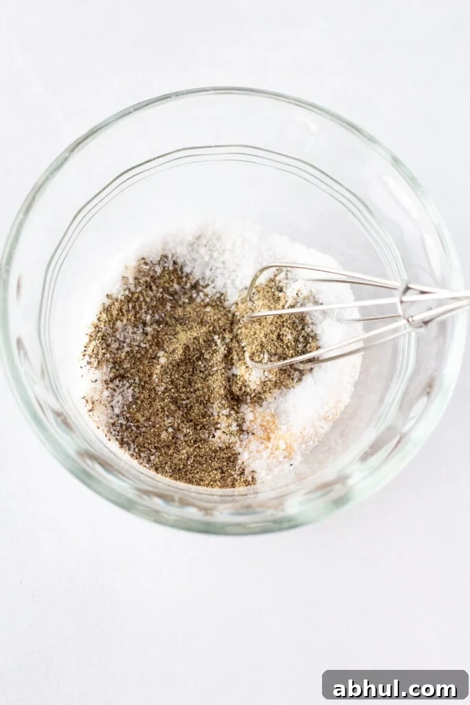 Close up of a small whisk stirring ground black pepper, kosher salt, and garlic powder together in a small clear bowl that rests on a white surface.