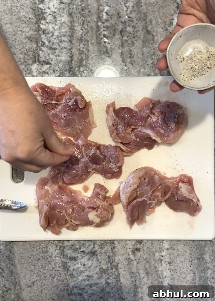 Top down shot of a hand seasoning raw boneless skinless chicken thighs on a white cutting board.
