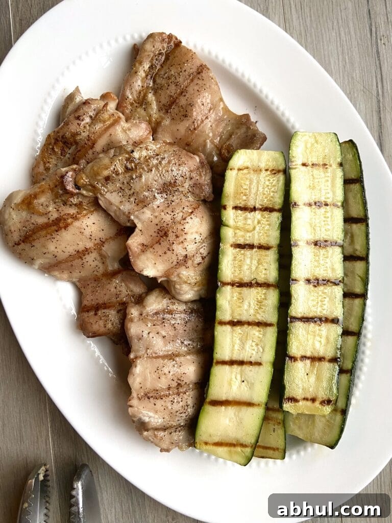 Top down shot of grilled boneless skinless chicken thighs next to grilled zucchini on a white platter.