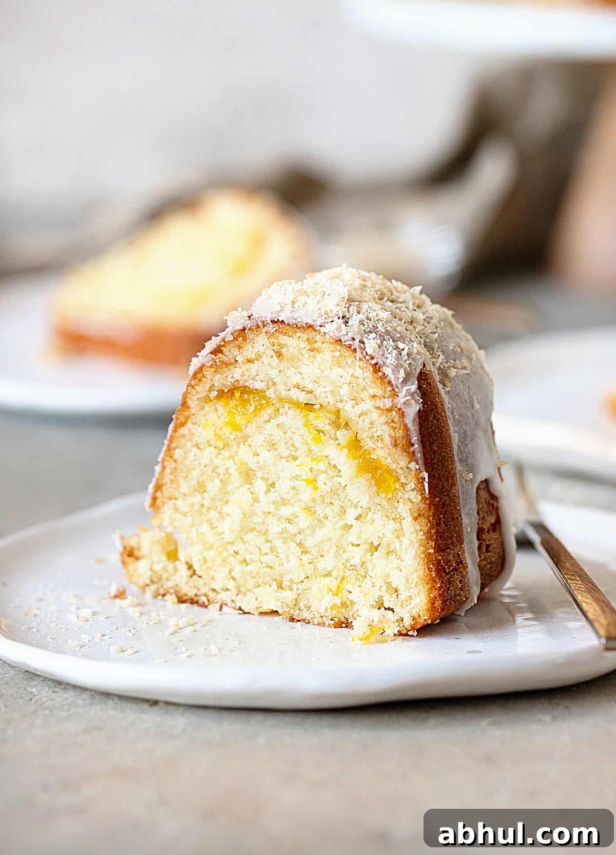 Moist Mango Coconut Bundt Cake Recipe for Tropical Desserts 2 Slice of mango bundt cake on a white plate. Grey surface and background with more slices.