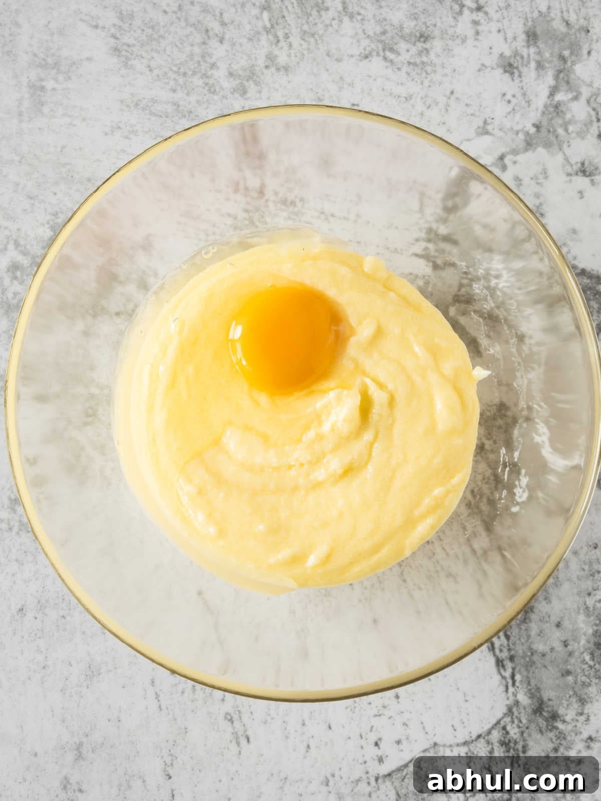 Moist Mango Coconut Bundt Cake Recipe for Tropical Desserts 6 Egg added to glass bowl with cake batter on a grey surface. Top view.