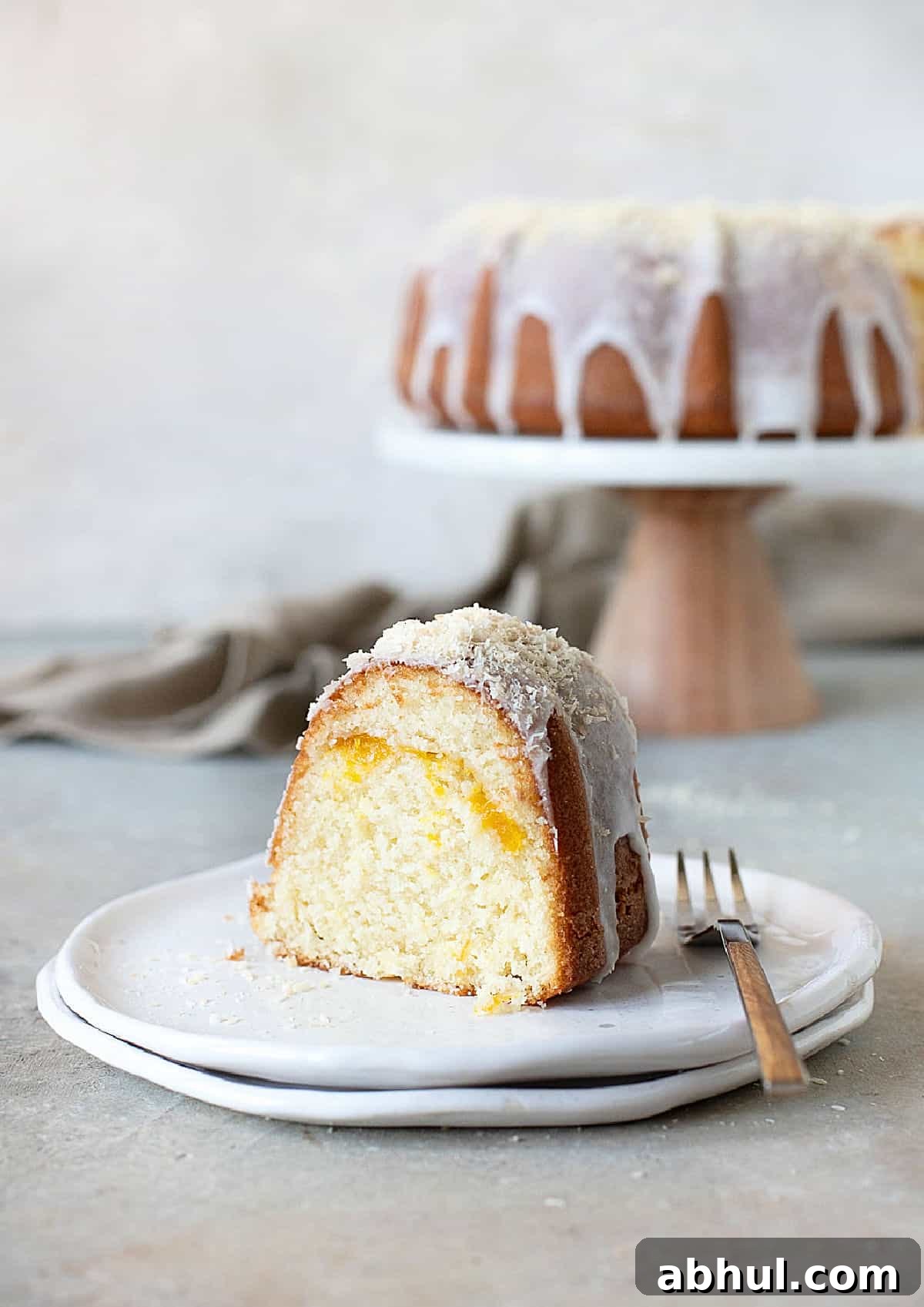 Moist Mango Coconut Bundt Cake Recipe for Tropical Desserts 9 Glazed mango coconut bundt cake on a white plate. Cake stand in the grey background. Grey surface.