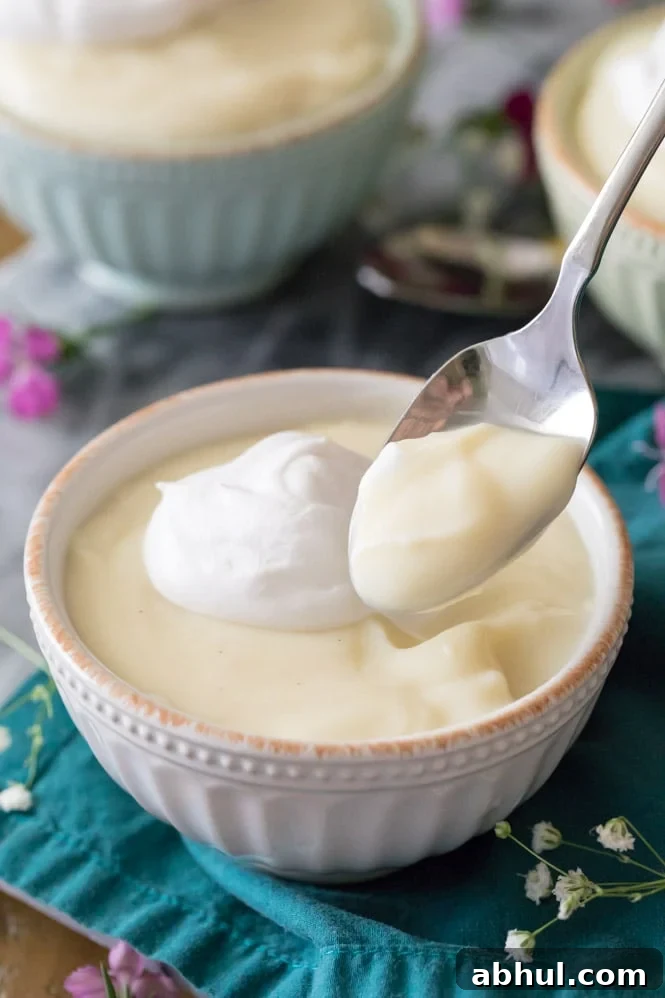 Spoon with vanilla pudding on it, bowl of vanilla pudding topped with whipped cream in the background