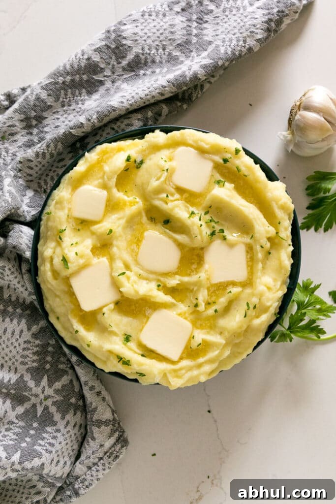 An overhead image of a bowl of mashed potatoes with melting pats of butter and chopped parsley on top with a head of garlic, grey napkin and sprigs of parsley round it.
