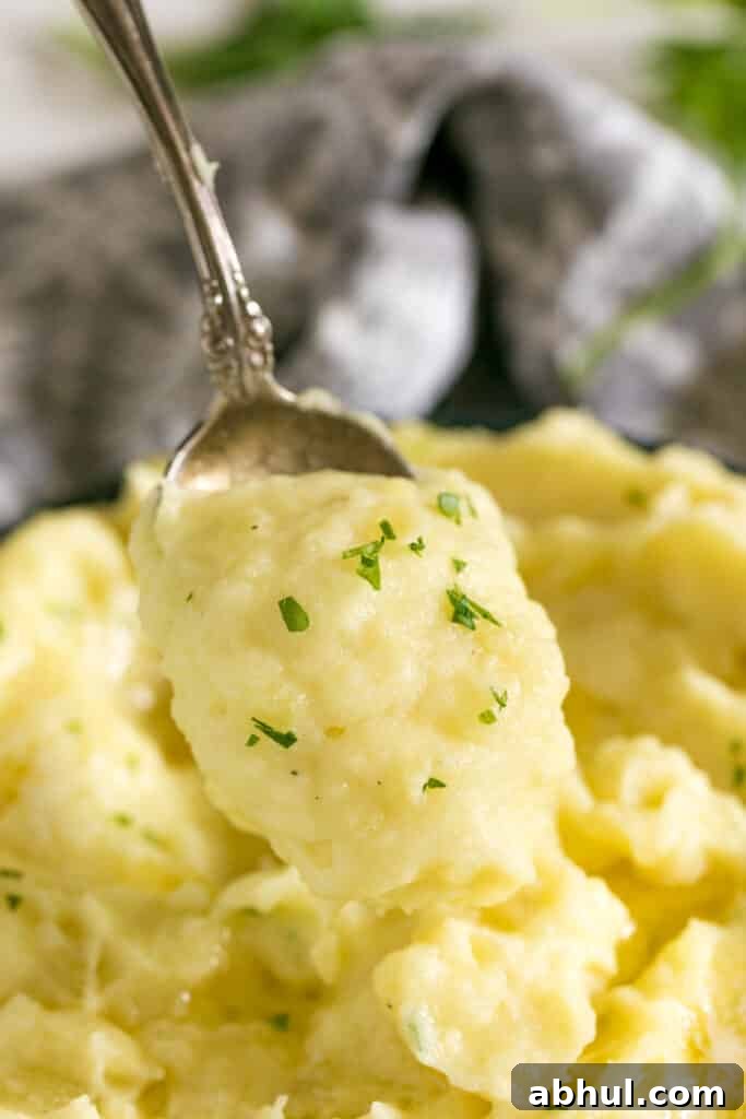 A spoon holding a scoop of mashed potatoes with a bowl of them behind it and a grey napkin.