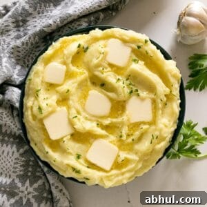 An overhead image of a bowl of mashed potatoes with melting pats of butter and chopped parsley on top with a head of garlic, grey napkin and sprigs of parsley round it.