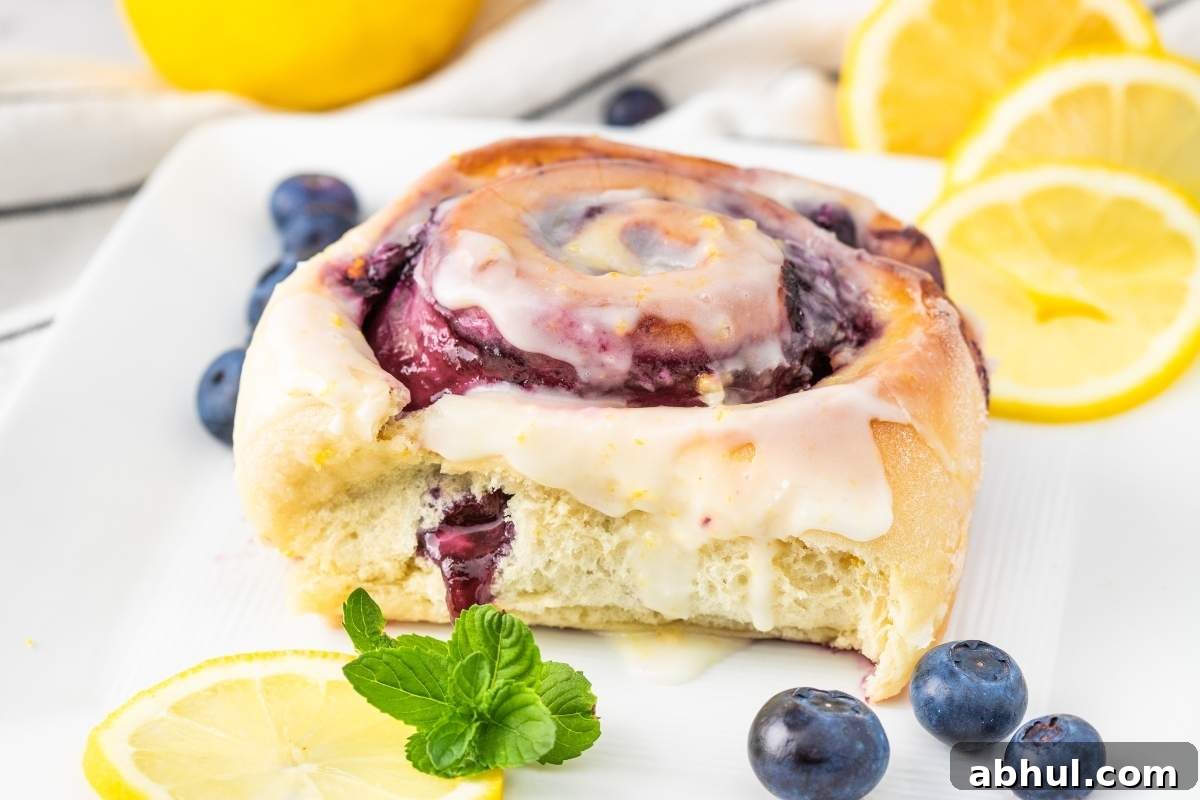 A lemon blueberry sweet roll with icing, garnished with fresh blueberries and lemon slices, sits invitingly on a white plate.