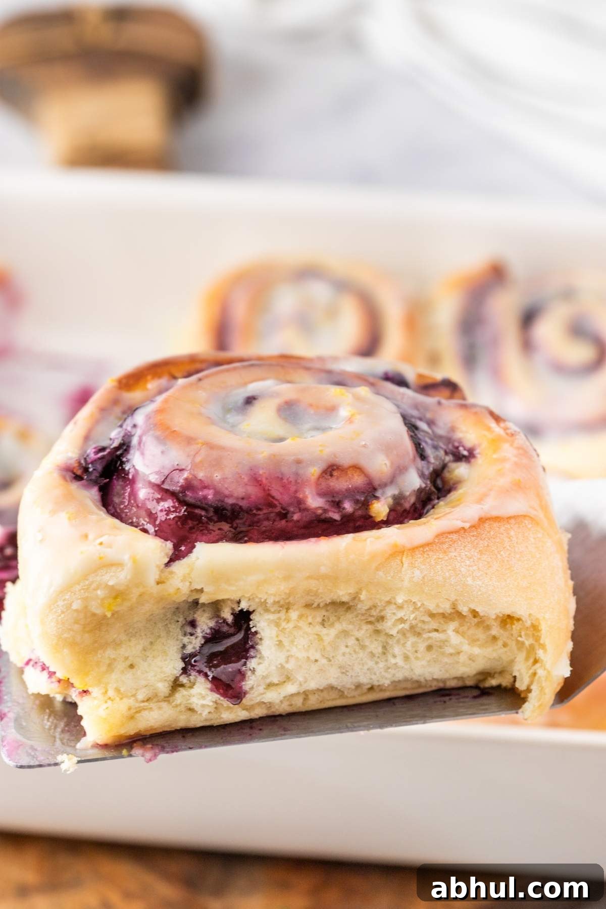 Close-up of a glazed cinnamon roll with purple blueberry filling and a hint of lemon zest, featuring a bite taken out. The roll is held above a baking dish filled with more delicious sweet rolls.