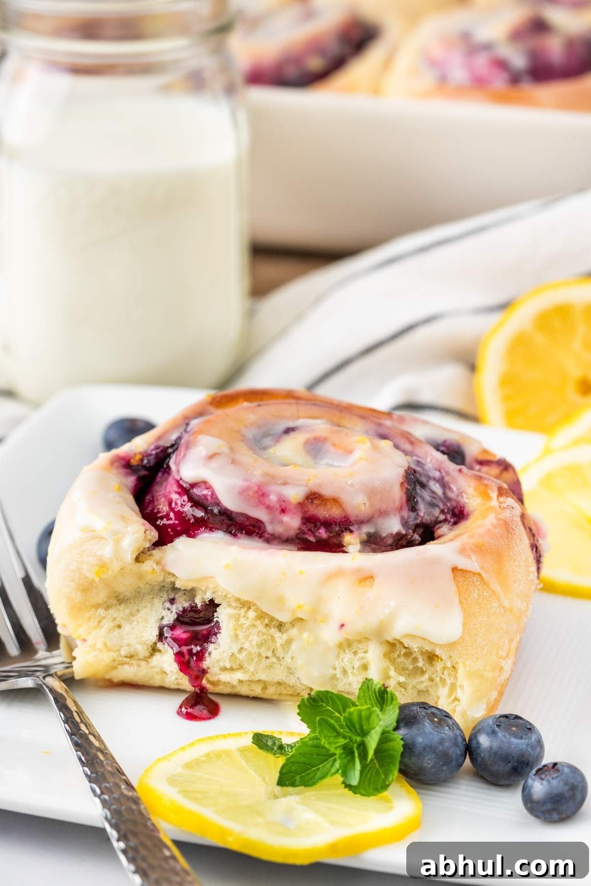 A lemon blueberry sweet roll with icing sits elegantly on a plate, adorned with fresh blueberries, lemon slices, and mint. Nearby, a fork lies ready beside a refreshing glass of milk.