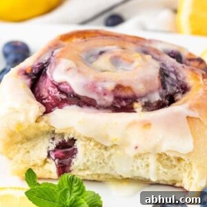 A glazed lemon blueberry sweet roll with berry filling sits elegantly on a white plate, garnished with fresh lemon slices, juicy blueberries, and sprigs of mint.