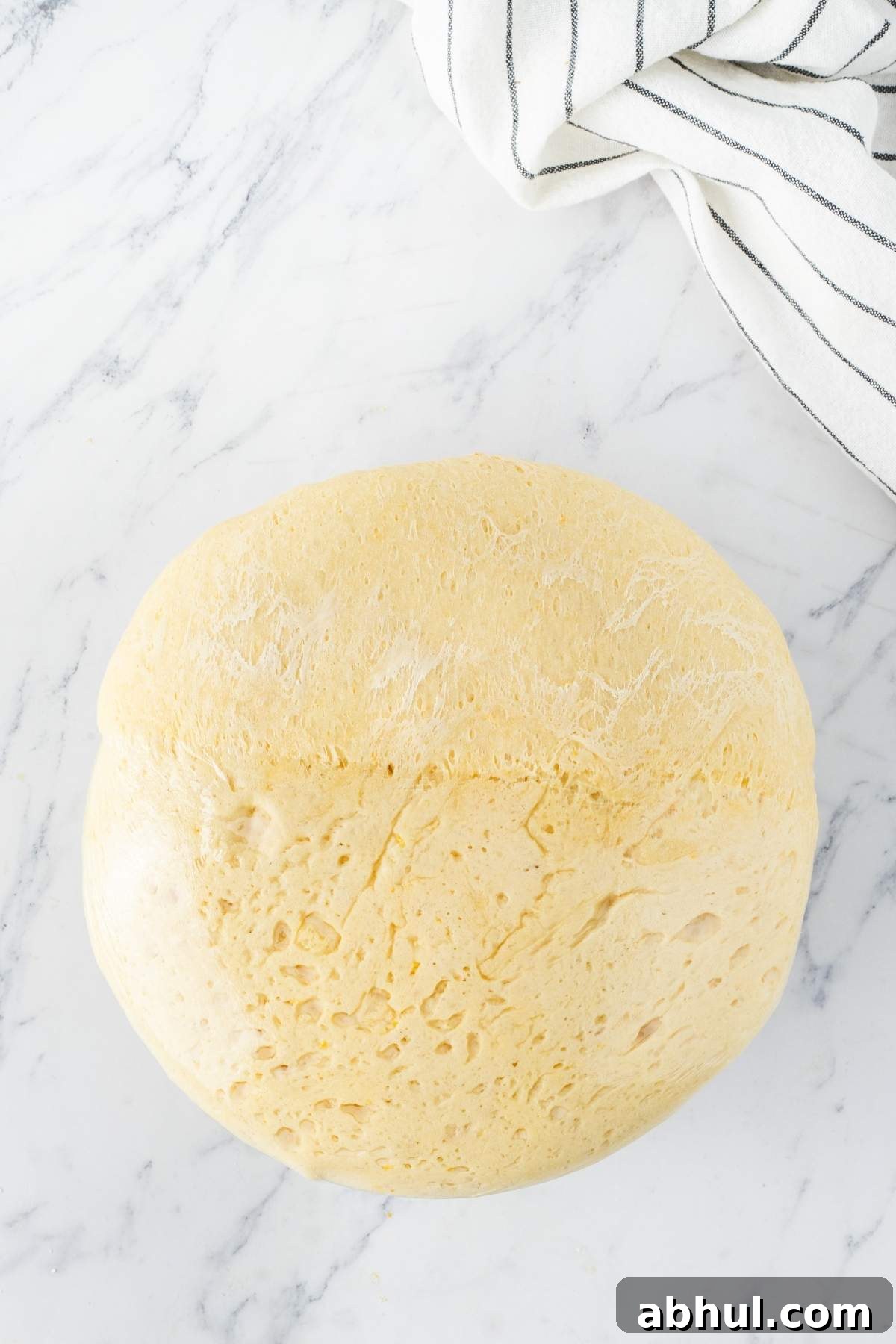 A ball of risen dough on a marble surface, poised to become delicious lemon blueberry sweet rolls, rests beside a white cloth with black stripes.