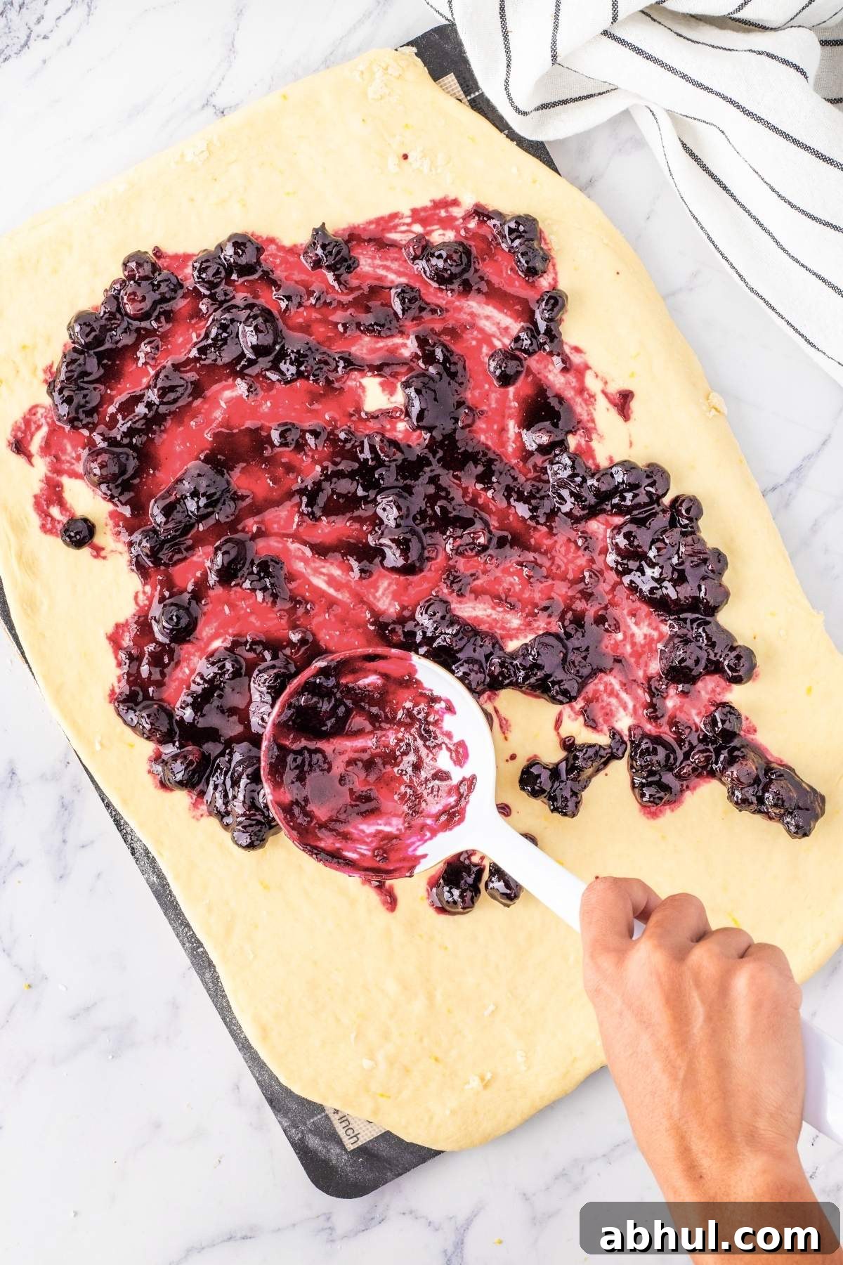 A hand spreads blueberry filling on rolled-out dough with a white spatula on a marble surface, preparing the base for delightful lemon blueberry sweet rolls.