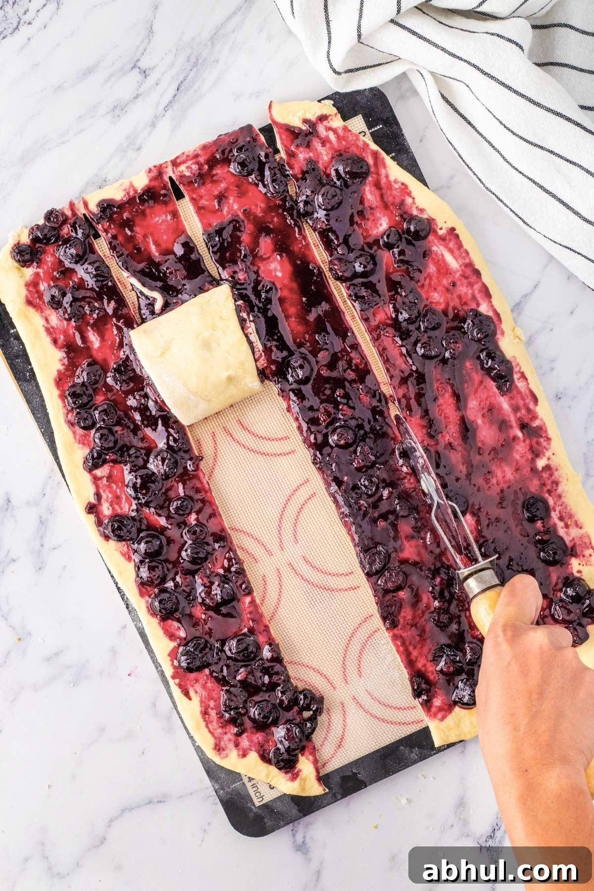 A person skillfully cuts cherry-filled pastry dough into strips on a marble surface, with a striped cloth nearby, dreaming of crafting delightful lemon blueberry sweet rolls.