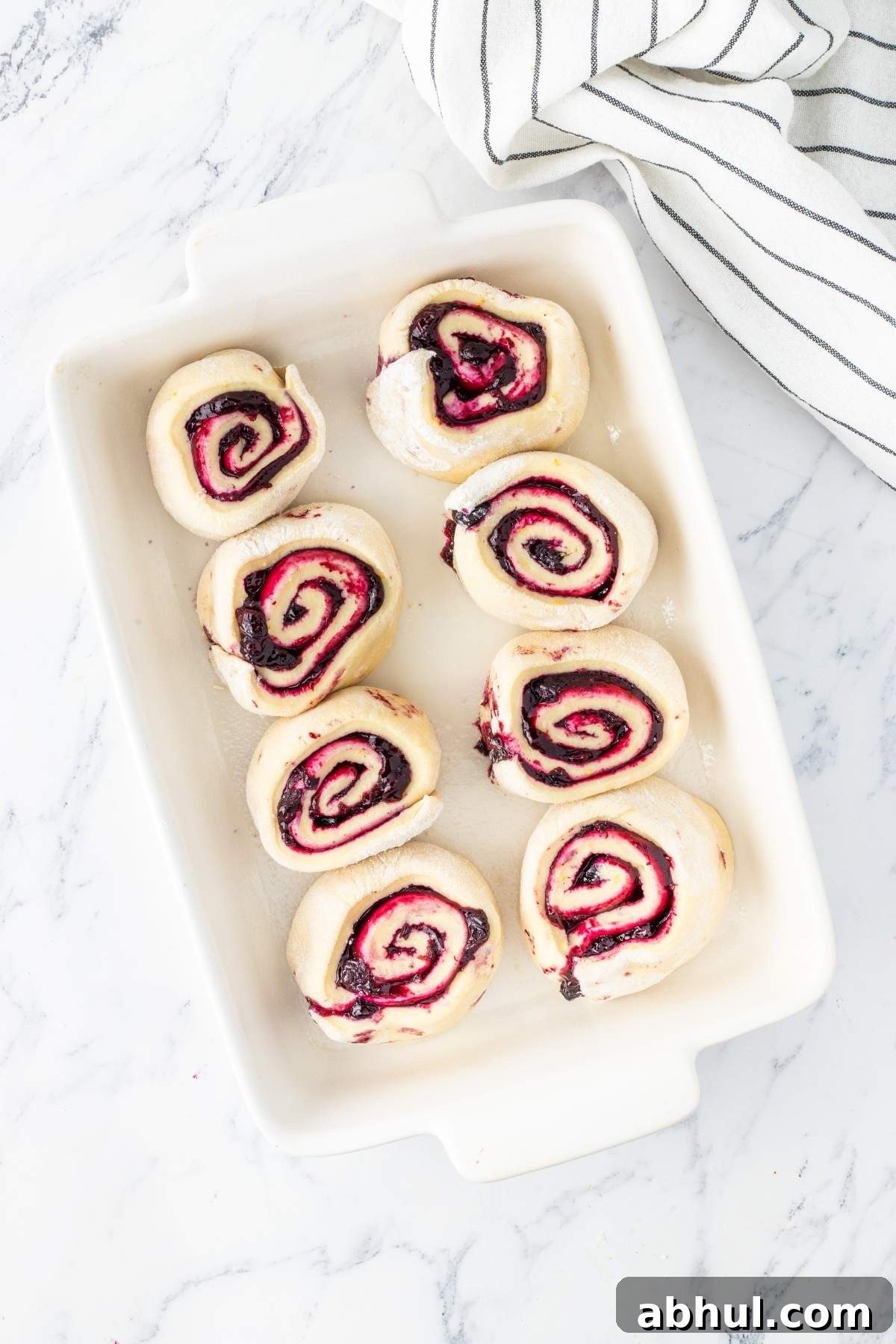 Eight uncooked lemon blueberry sweet rolls in a white rectangular baking dish on a marble surface, with a striped cloth nearby.