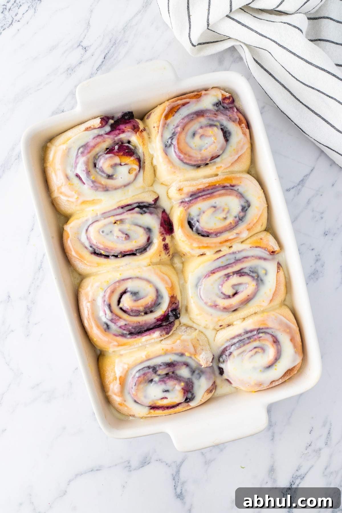 A baking dish with eight frosted cinnamon rolls, infused with a hint of lemon blueberry sweetness, arranged in two rows on a marble surface. A striped cloth is partially visible in the top right corner.
