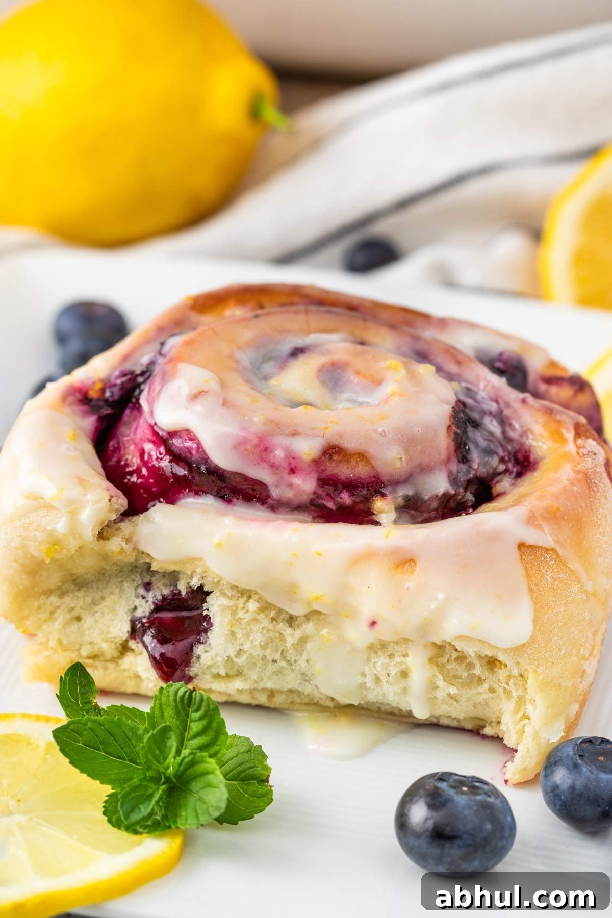 Close-up of lemon blueberry sweet rolls drizzled with white icing, surrounded by fresh blueberries, a lemon slice, and a sprig of mint on a white plate.
