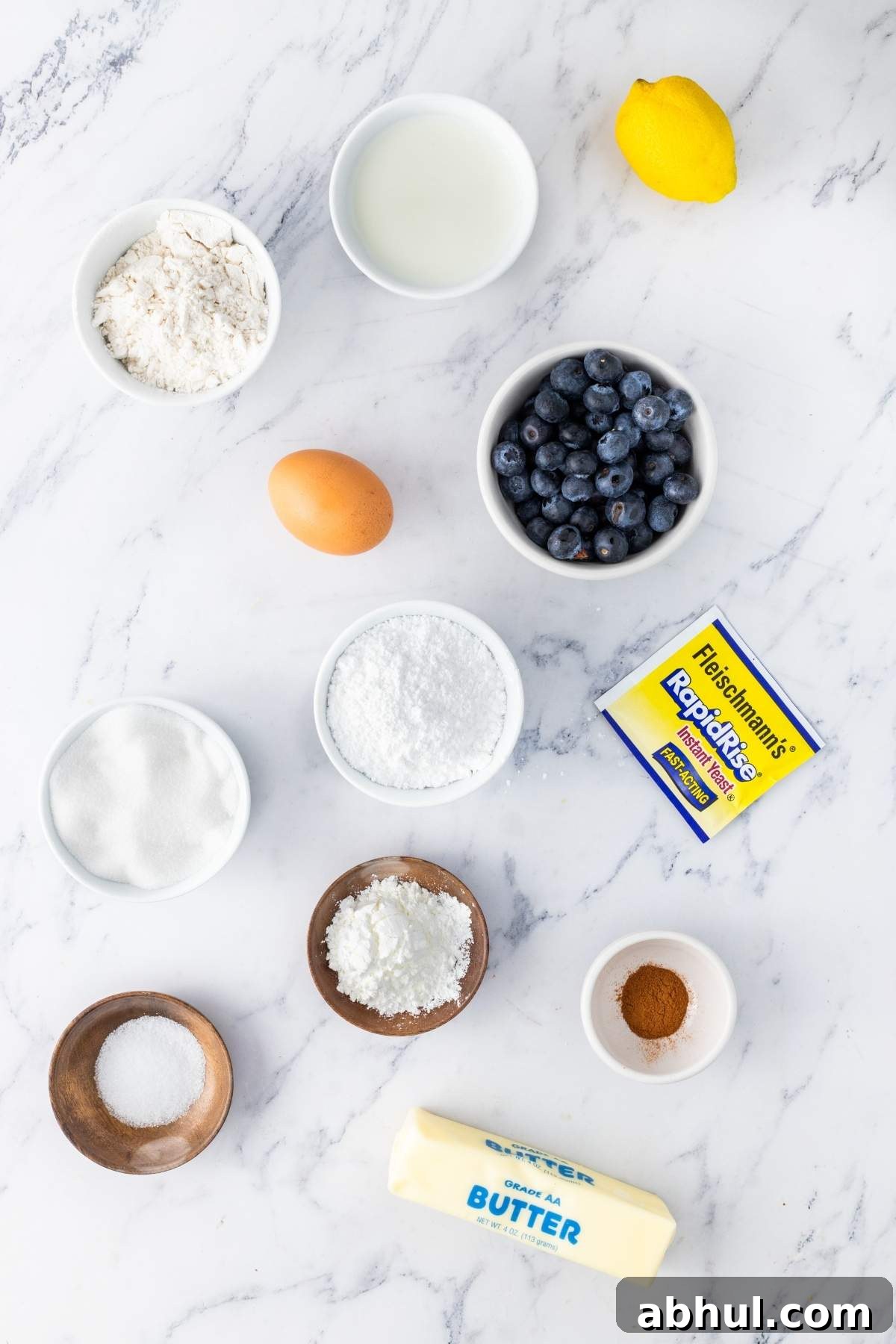 Ingredients for baking lemon blueberry sweet rolls: flour, egg, milk, lemon, blueberries, butter, sugar, yeast, baking powder, salt, and cinnamon scattered on a marble surface.