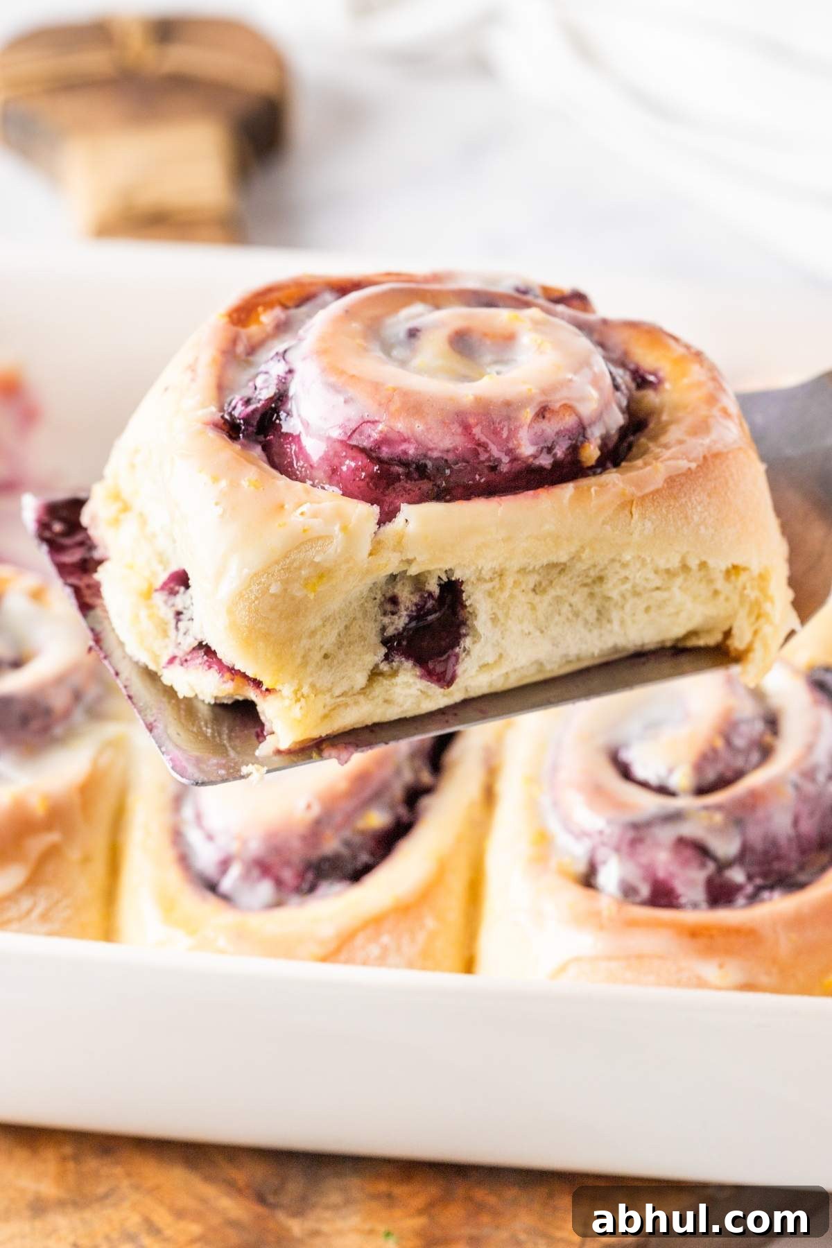 A close-up of a freshly baked lemon blueberry sweet roll with icing being lifted from a baking dish using a spatula.