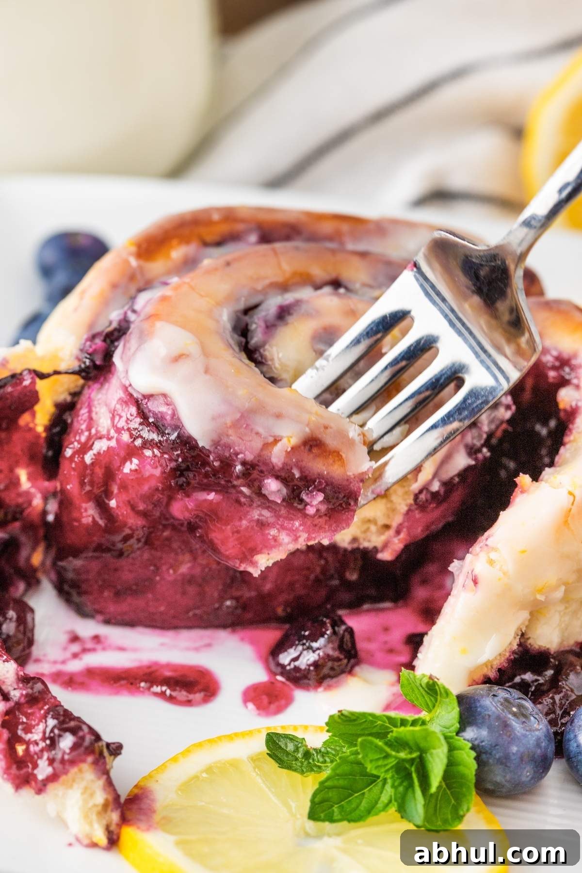 Close-up of a partially eaten glazed lemon blueberry sweet roll on a plate, accompanied by a fork. A fresh lemon slice, scattered blueberries, and mint leaves elegantly garnish the plate.