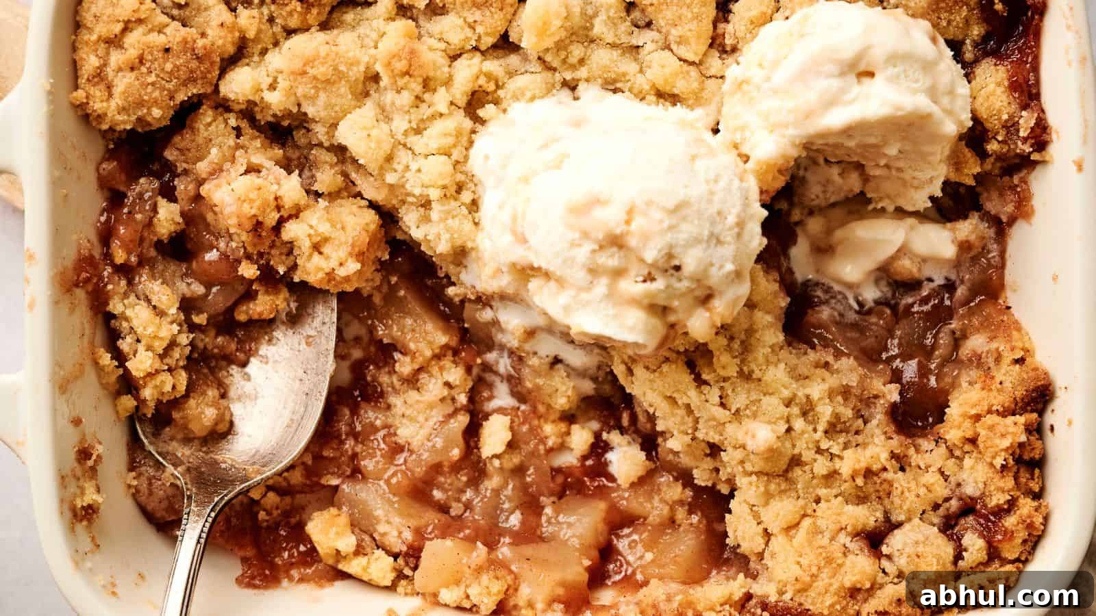 Close-up of an apple crumble dessert with a golden crust, topped with two scoops of vanilla ice cream, and a spoon placed inside the dish.