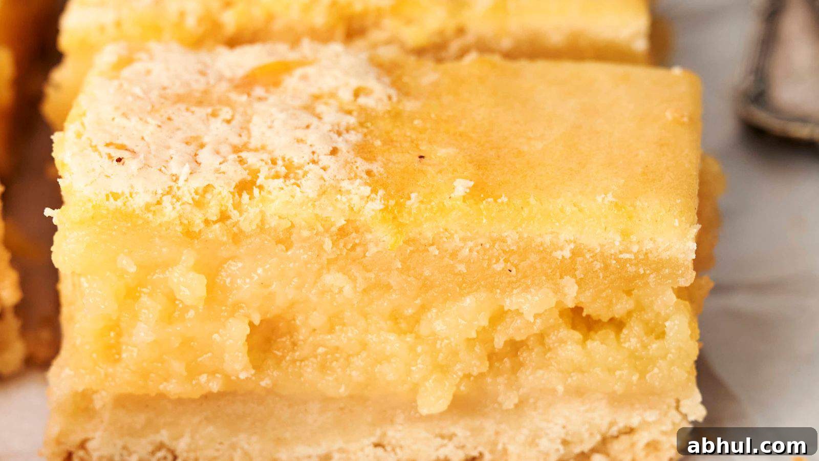 Close-up of a lemon bar with a crumbly, light yellow top layer and a thicker, moist filling resting on a shortbread crust.